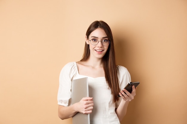 Sorridente giovane donna con gli occhiali leggendo notizie emozionanti sul cellulare, tenendo il laptop e guardando felice la fotocamera, in piedi su sfondo beige.