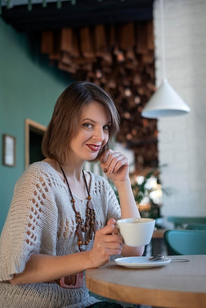Sorridente giovane donna in abito grigio beve caffè all'interno del bar ritratto di ragazza bruna si siede al tavolo in caffè