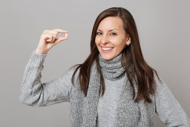 Smiling young woman in gray sweater, scarf holding medication tablet, aspirin pill isolated on grey background. Healthy lifestyle, ill sick disease treatment, cold season concept. Mock up copy space.