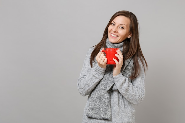 Smiling young woman in gray sweater, scarf holding red cup of coffee or tea isolated on grey wall background. Healthy fashion lifestyle people sincere emotions cold season concept. Mock up copy space.