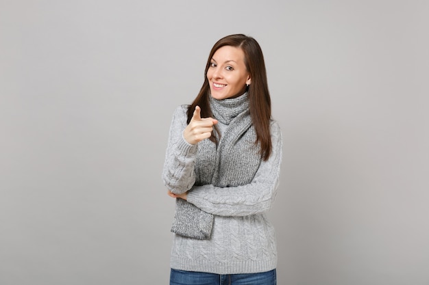 灰色のセーター、灰色の壁の背景に分離されたカメラに人差し指を指すスカーフで若い女性の笑顔。健康的なファッションライフスタイル、人々の誠実な感情の寒い季節のコンセプト。コピースペースをモックアップします。
