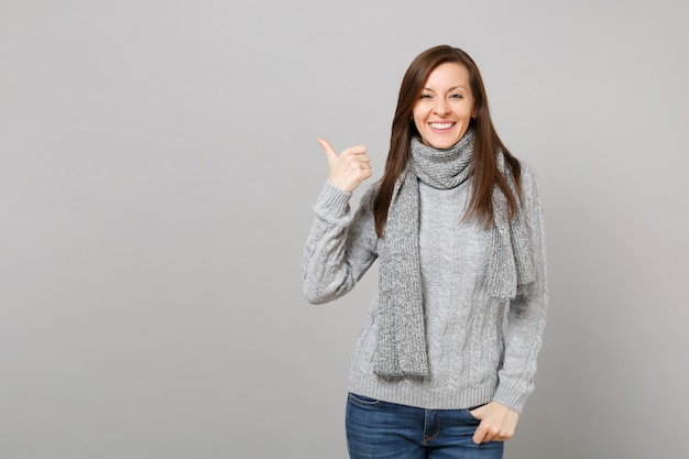 Smiling young woman in gray sweater, scarf pointing thumb aside isolated on grey wall background in studio. Healthy fashion lifestyle, people sincere emotions, cold season concept. Mock up copy space.