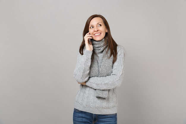 회색 스웨터를 입은 웃고 있는 젊은 여성, 스카프는 휴대전화로 통화하고, 회색 벽 배경에서 격리된 즐거운 대화를 합니다. 건강한 패션 라이프 스타일, 사람들의 감정, 추운 계절 개념.
