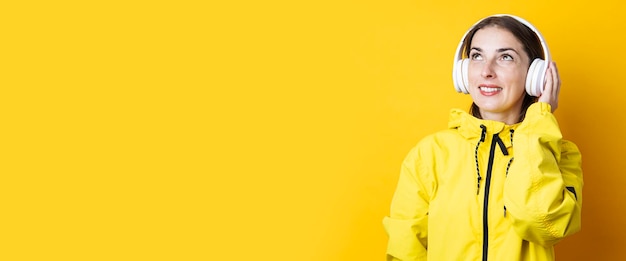 Smiling young woman in headphones in a yellow jacket looks up on a yellow background Banner