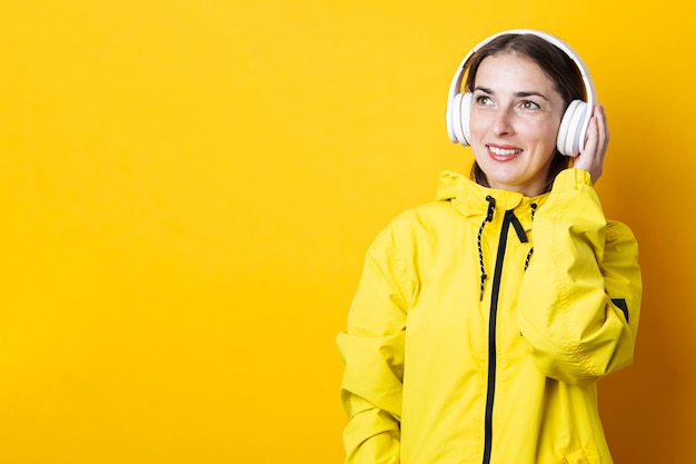 Smiling young woman in headphones in a yellow jacket looks up on a yellow background