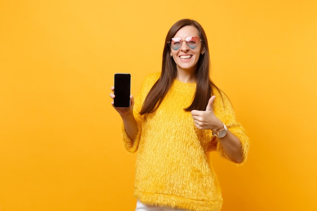 Smiling young woman in heart glasses showing thumb up and holding mobile phone with blank black empty screen isolated on bright yellow background. People sincere emotions, lifestyle. Advertising area.