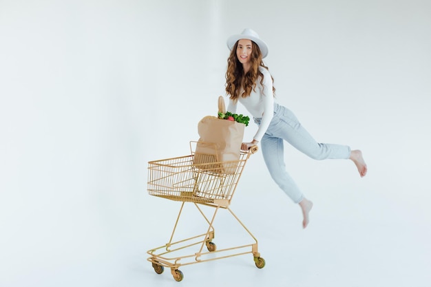 smiling young woman holding apple and looking away while standing with shopping trolley