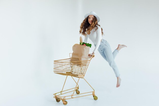 smiling young woman holding apple and looking away while standing with shopping trolley