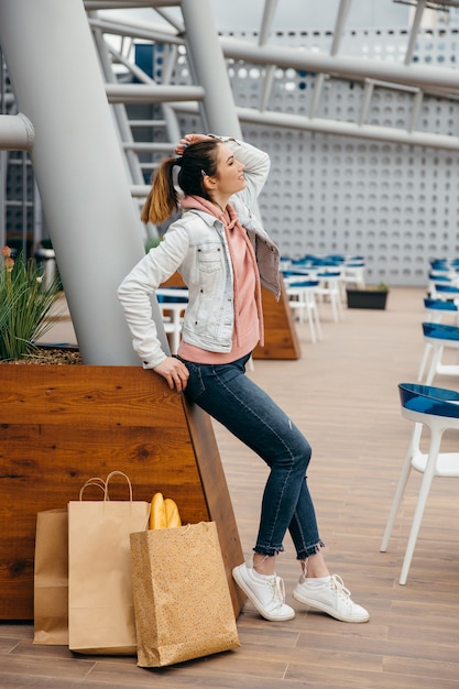 Smiling young woman holding coffee cup, walking with grocery shopping paper bag with long white bread baguette