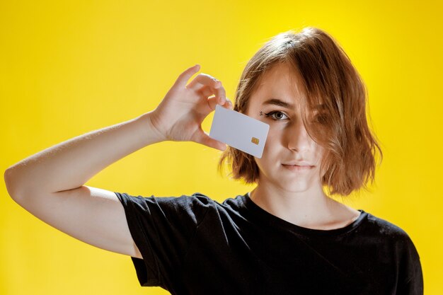 Smiling young woman holding credit card on yellow background.