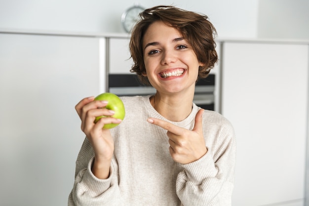 Smiling young woman holding green apple at the kitchen