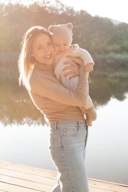 Smiling young woman holding playing with baby boy 1 year old wear knit clothes over nature background and lake with forest Autumn season Motherhood