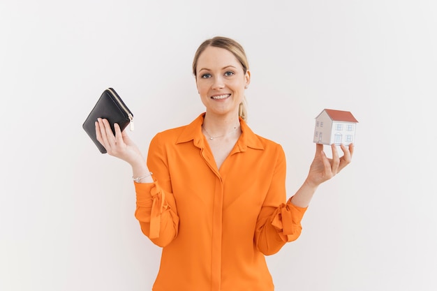 Smiling young woman holding wallet and miniature house model isolated on white wall