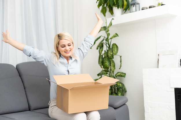 Sorridente giovane donna a casa sul divano, ha ricevuto un pacco postale, shopping online e concetto di consegna