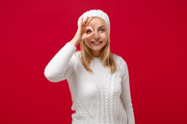 写真 白いセーターを着た笑顔の若い女性、スタジオで赤い背景に隔離された帽子。健康的なファッション ライフ スタイル、寒い季節のコンセプト。コピー スペースをモックアップします。双眼鏡や眼鏡の真似をして ok のジェスチャーを示します。