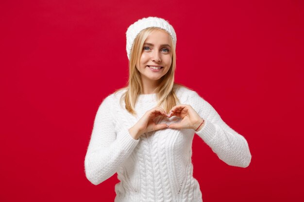 Фото Улыбающаяся молодая женщина в белом свитере, шляпе на красном фоне в студии. здоровый модный образ жизни, концепция холодного сезона. скопируйте пространство для копирования. показывает форму сердца руками, знак в форме сердца.