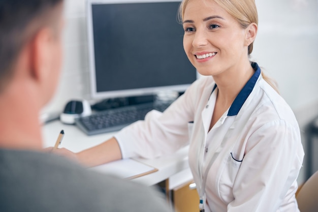 Smiling young woman is listening to male patient while sitting at desk with computer in office