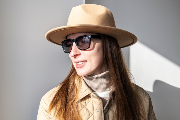 Smiling young woman in a jacket in a hat in sunglasses against a white wall