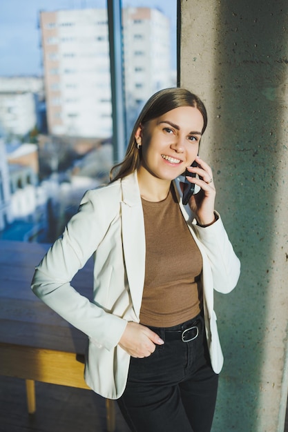 A smiling young woman in a jacket stands in the office and happily talks on the phone A young manager works in the office and manages the business remotely