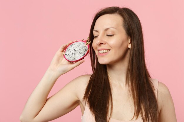 Giovane donna sorridente che tiene gli occhi chiusi tenendo pitahaya fresco maturo, frutto del drago isolato su sfondo rosa pastello in studio. lo stile di vita vivido delle persone si rilassa il concetto di vacanza. mock up spazio di copia.