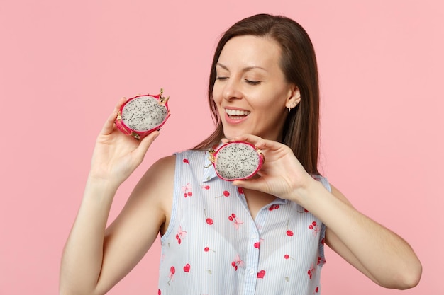 Giovane donna sorridente che tiene gli occhi chiusi tenendo metà di pitahaya fresco maturo, frutto del drago isolato su sfondo rosa pastello. lo stile di vita vivido delle persone si rilassa il concetto di vacanza. mock up spazio di copia.