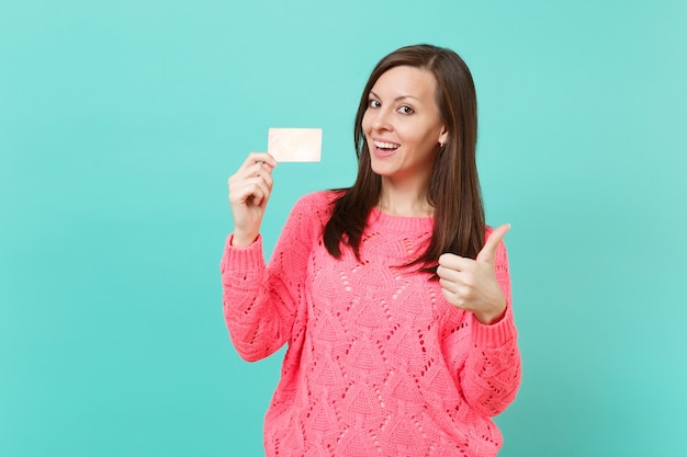 Smiling young woman in knitted pink sweater showing thumb up and holding credit card in hand isolated on blue turquoise wall background studio portrait. People lifestyle concept. Mock up copy space.