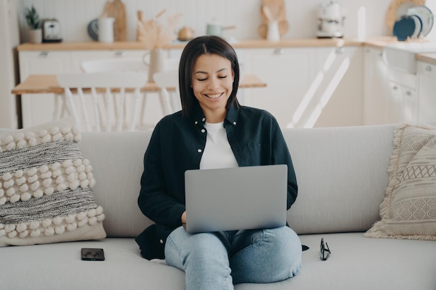 Smiling young woman learning online at laptop sitting on sofa at home Distance education elearning