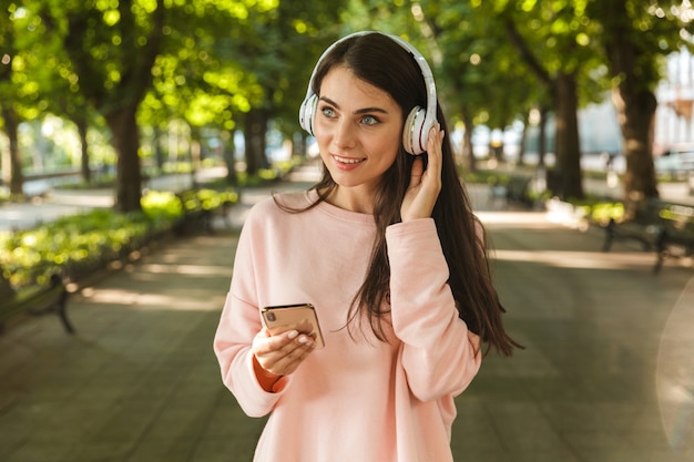 Smiling young woman listening to music with headphones while walking at the city park and holding mobile phone