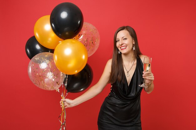 Smiling young woman in little black dress celebrating, holding glass of champagne, air balloons isolated on red background. St. Valentine's Day, Happy New Year, birthday mockup holiday party concept.