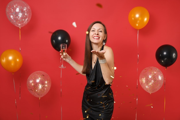 Smiling young woman in little black dress celebrating, holding glass of champagne and catching confetti on bright red background air balloons. Happy New Year, birthday mockup holiday party concept.