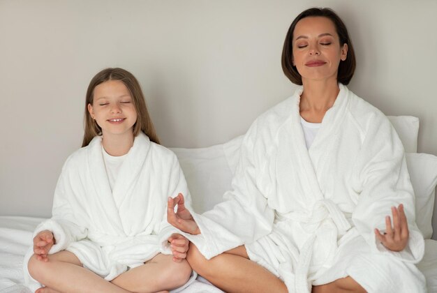 Smiling young woman and little caucasian girl in bathrobe with closed eyes meditation resting on white bed