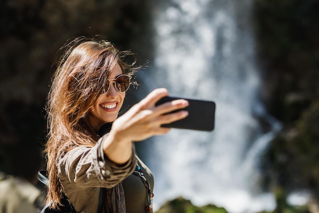 아름다운 폭포의 전망을 즐기면서 스마트폰으로 셀카를 만드는 웃고 있는 젊은 여성.