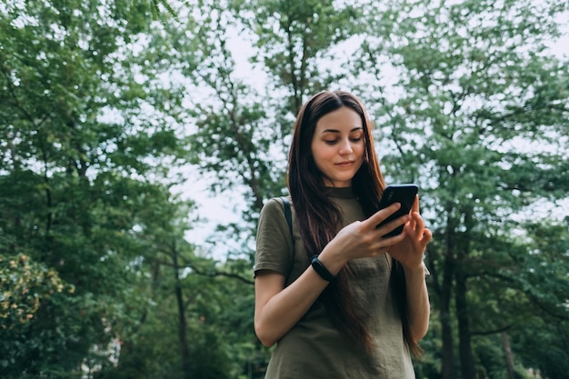 Smiling young woman in the park, using smartphone and typing text.