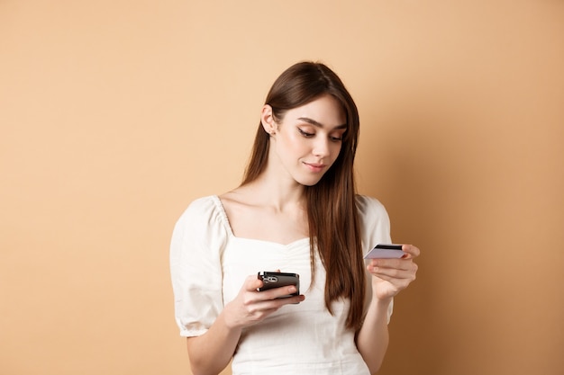 Smiling young woman paying online, looking at credit card and making purchase on mobile phone, shopping in internet, standing on beige.