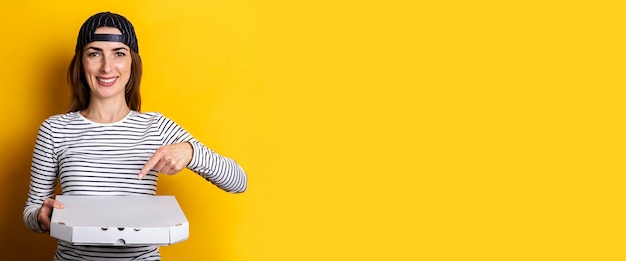 Smiling young woman pizza delivery man points finger at holding pizza on yellow background Banner