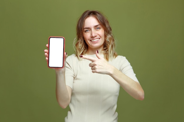 Smiling young woman pointing by finger on phone white blank mock up screen happy with mobile apps