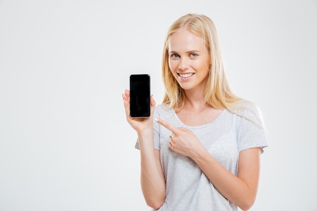 Smiling young woman pointing finger at blank smartphone screen isolated on a white wall