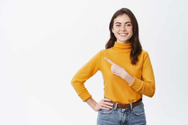 Smiling young woman pointing finger left at banner, showing advertisement on white wall