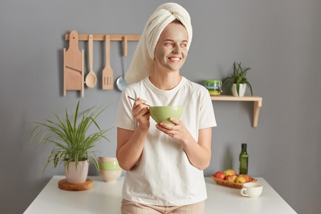 Smiling young woman posing in kitchen with towel on head having breakfast standing with plate in hands doing cosmetics procedures in the morning