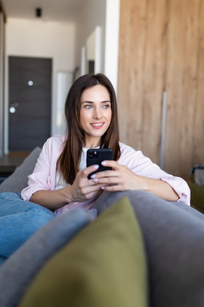 Smiling young woman reading sms while sitting on sofa at home