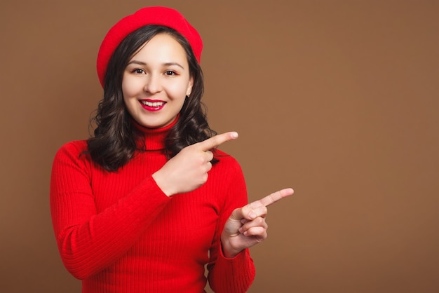 Smiling young woman in red beret and sweater shows gesture on empty beige