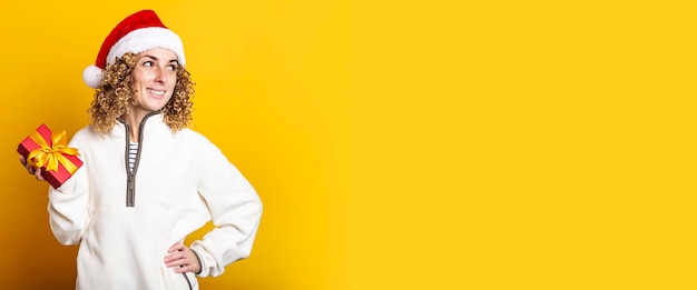 Smiling young woman in santa claus hat holding a gift on a yellow background. Banner.