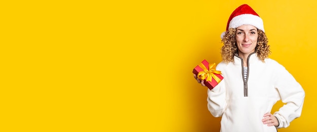 Smiling young woman in santa claus hat holding a gift on a yellow background banner