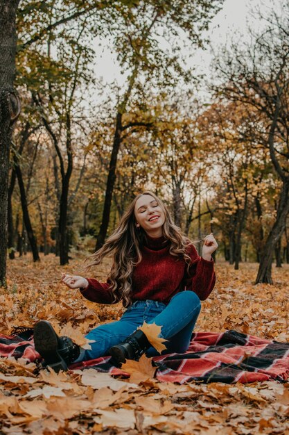 Улыбающаяся молодая женщина сидит на осенних листьях на деревьях в лесу