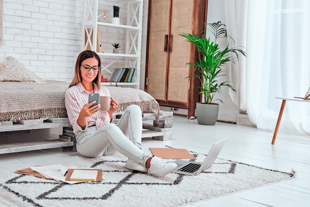 Smiling young woman sitting on floor at home with cell phone and drinking coffee or tea Copy space