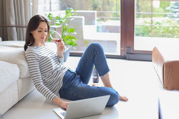 Smiling young woman sitting on floor with laptop computer and chating with friends drinking wine