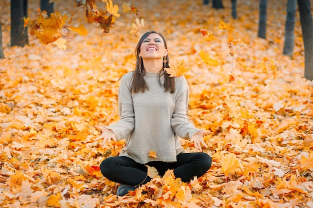 Foto la giovane donna sorridente che si siede sulle foglie che si diverte nel parco di autunno solleva le foglie cadute