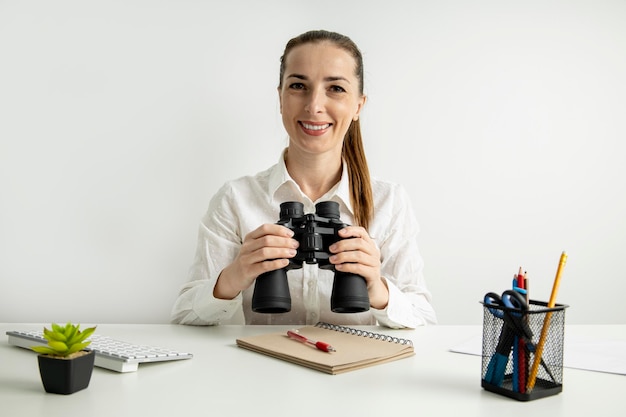 Smiling young woman sitting in the office at her desk holding binoculars