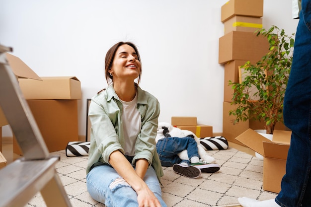 Улыбающаяся молодая женщина, сидящая с коробками на ковре, переезжает в новую квартиру
