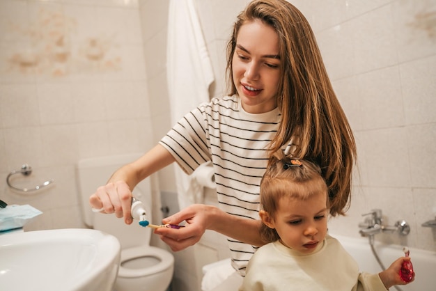 Foto sorridente giovane donna che spreme il dentifricio dal tubo sullo spazzolino del suo bambino